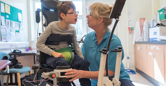 A smiling boy in the Innowalk Pro, a motorized standing exercise device, is assisted by a therapist.